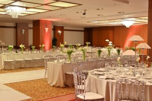 Geniş düğün salonunda beyaz çiçekli yuvarlak masalar, kristal sandalyeler, avizeler ve renkli duvar aydınlatmasıyla hazırlanmış genel salon düzeni